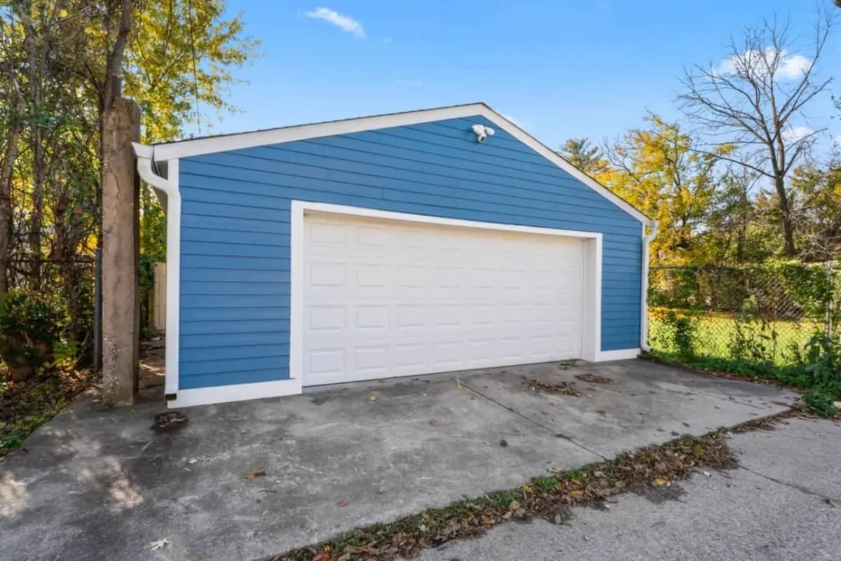 This shot showcases a blue garage with a crisp white door, framed by lush greenery. The space feels functional and approachable, suggesting ample room for parking or even small outdoor gatherings adjacent to the home.