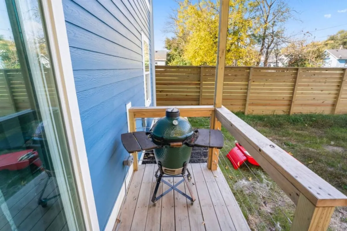 A welcoming wooden deck accented by a green grill and the home's blue exterior wall. The space is arranged for outdoor cooking, social gatherings, and casual relaxation in the sun, suggesting a natural flow from the hot tub area to other outdoor amenities.