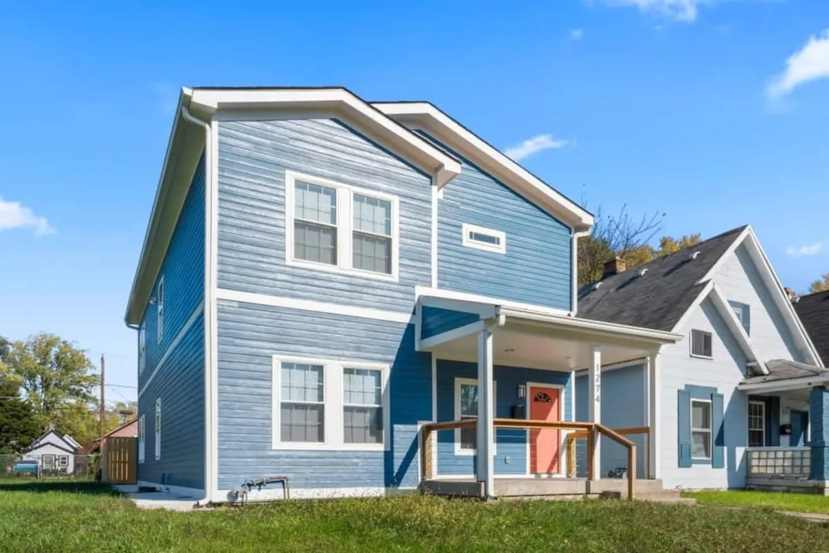 The image displays the home's bright blue siding, an inviting orange front door, and a cozy front porch surrounded by greenery. The cheerful exterior hints at a welcoming atmosphere and the possibility of enjoying time outdoors with loved ones, whether relaxing on the porch or gathering in the yard.