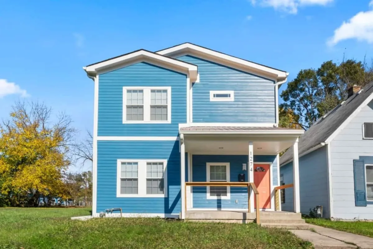 This image features the blue exterior of the home, highlighted by a welcoming red front door, an inviting porch area, and lush surrounding greenery. The scene suggests a space perfect for family gatherings, outdoor play, and enjoying the outdoors together.