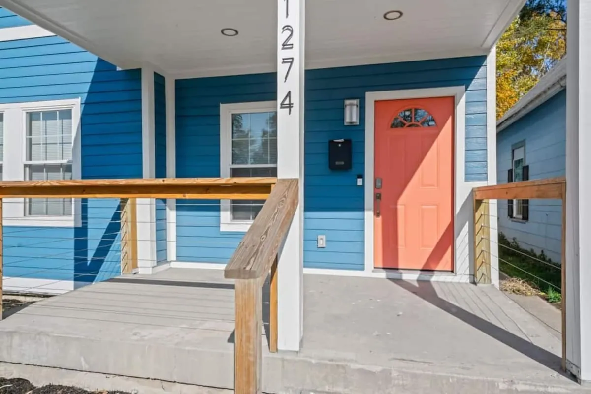 A close-up of the home's bright coral door set against blue siding and a wooden railing, capturing the inviting atmosphere at the entrance. The image hints at warm welcomes and joyful arrivals.