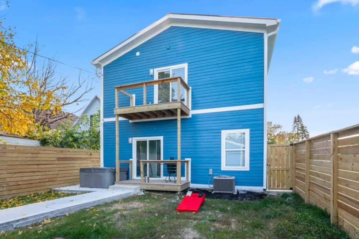 A bright and cheerful backyard scene showing the home's blue exterior, wooden balcony, and securely fenced yard. The setting is inviting for both peaceful solitude and group activities, emphasizing privacy and versatility in how the outdoor space can be enjoyed.