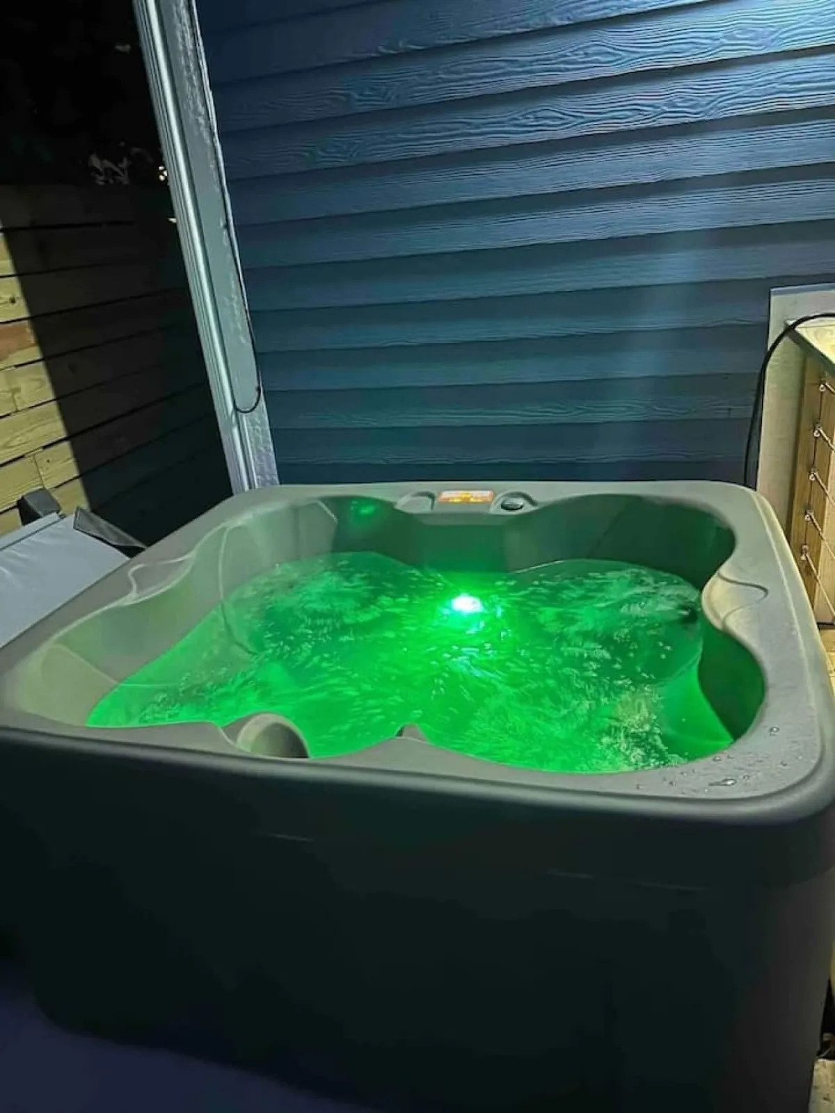 A tranquil outdoor scene centered on a private hot tub illuminated with a gentle green light, water gently swirling. The surrounding area hints at privacy and a cozy atmosphere perfect for small groups or intimate relaxation. The night sky above and subtle ambient lighting evoke a sense of unwinding under the stars.