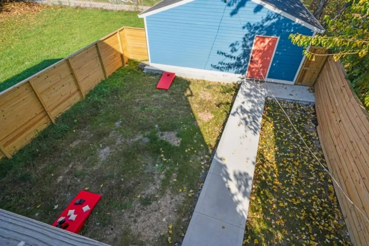 An outdoor fenced yard featuring a playful blue shed and bright red cornhole boards, ready for friendly games or summer parties. The scene exudes a fun, social vibe—perfect for group activities.