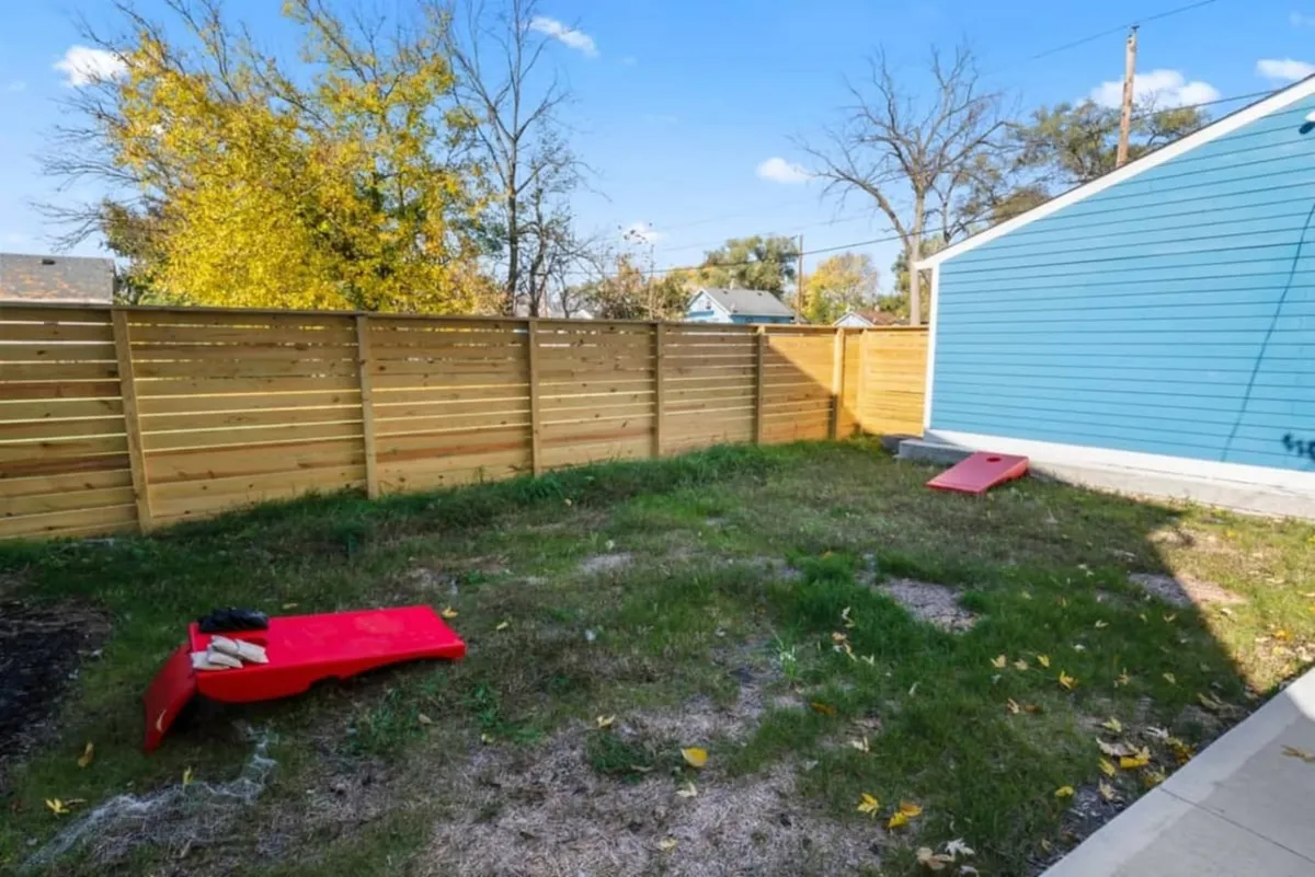 A playful backyard is shown with a wooden fence, blue exterior wall, green lawn, and vibrant red cornhole game boards. The space invites outdoor games, family gatherings, and relaxation in a sunny, cheerful setting.