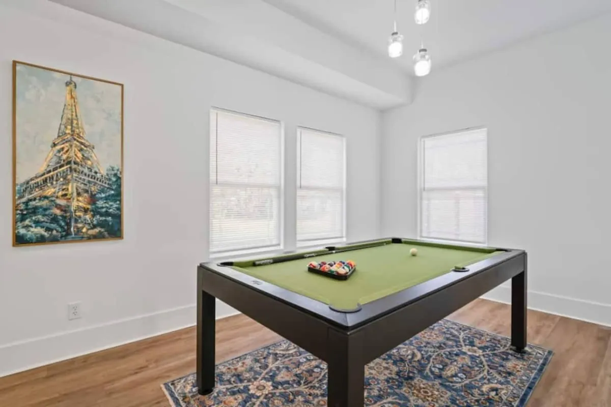 A bright interior game room centered around a pool table, with natural light streaming through the windows and artistic wall decor adding personality to the space. The setup encourages playful competition and casual hangouts.
