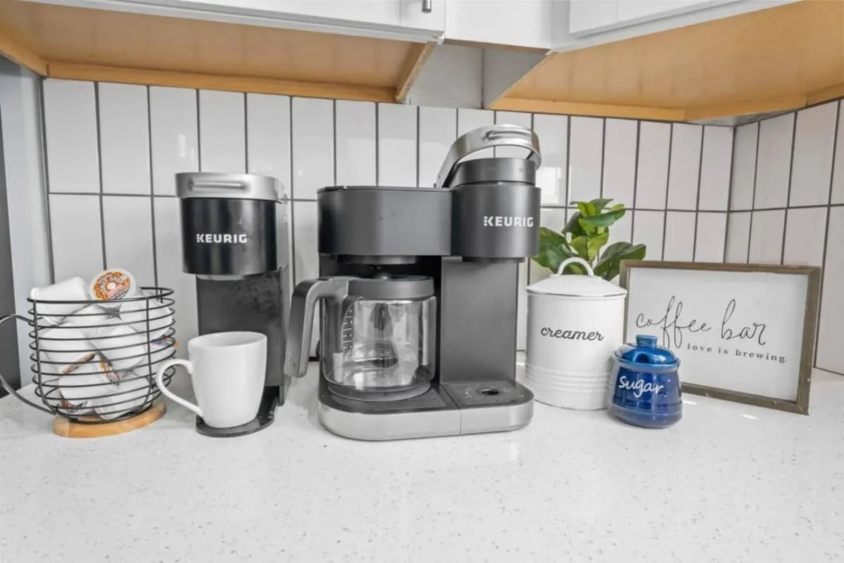 A close-up of a Keurig coffee maker, coffee mugs, and decorative containers arranged on a kitchen counter. This cozy vignette highlights the morning coffee ritual and the welcoming ambiance of the kitchen.
