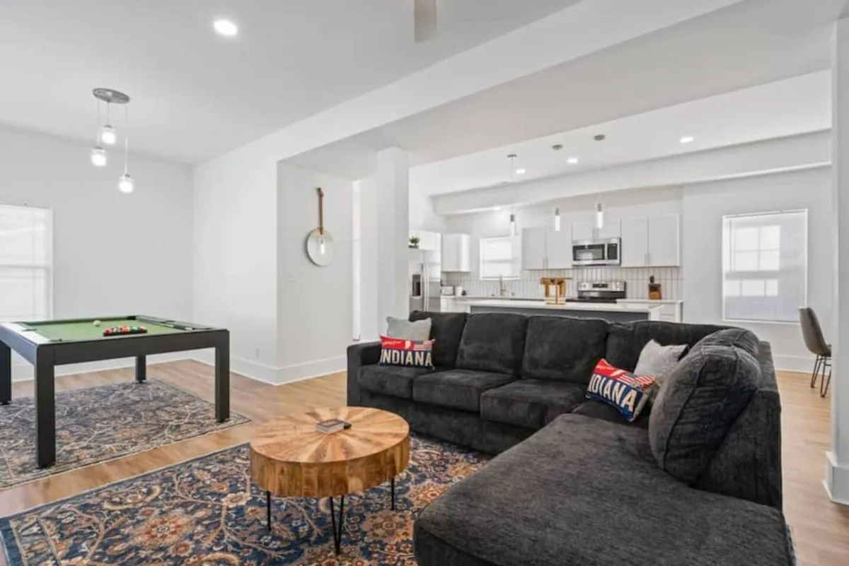 A spacious, open-concept living area that integrates a game room featuring a pool table, a comfortable sofa, a coffee table, and a glimpse of the kitchen area beyond. The space is filled with natural light and arranged to encourage social interaction and group activities.